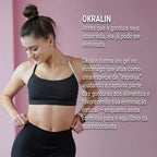 Dermafórmula Melhor Farmácia de Manipulação em Santos | Mulher jovem com coque no cabelo e roupa esportiva preta, olhando com satisfação para sua silhueta em um fundo rosa suave. O texto explica o diferencial do Okralin: a formação de um gel no estômago que captura gorduras dos alimentos antes da absorção, favorecendo a eliminação natural e o equilíbrio da microbiota.